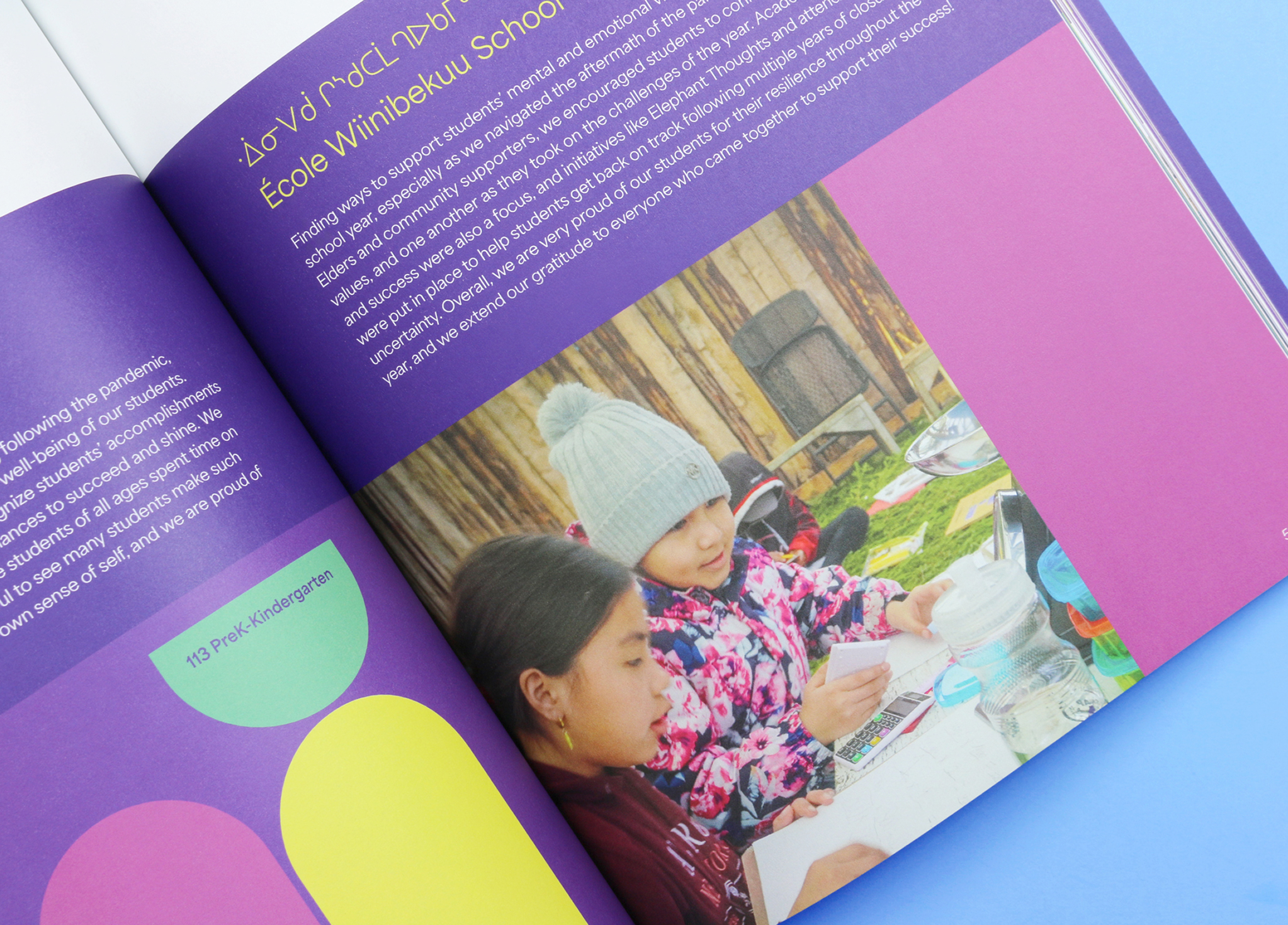 CSB_202223_Body-3 Two children sit at a table outdoors, writing in notebooks. One child wears a gray beanie and colorful jacket; the other has dark hair and a dark top. The scene appears in a colorful, open book with text in English and French above the image.