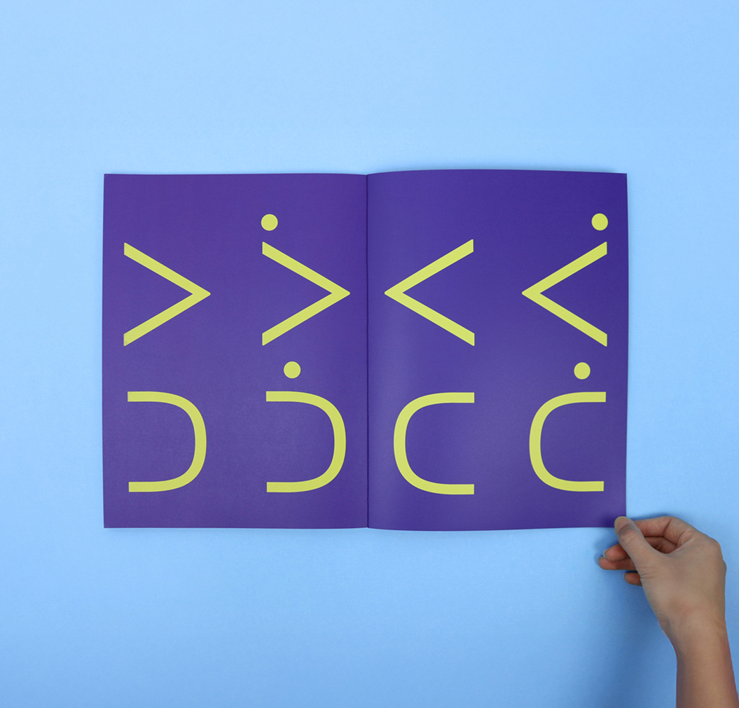 CSB_202223_Body-5 A hand holds open a book with a purple page, displaying abstract yellow shapes and symbols arranged in two rows against a light blue background.