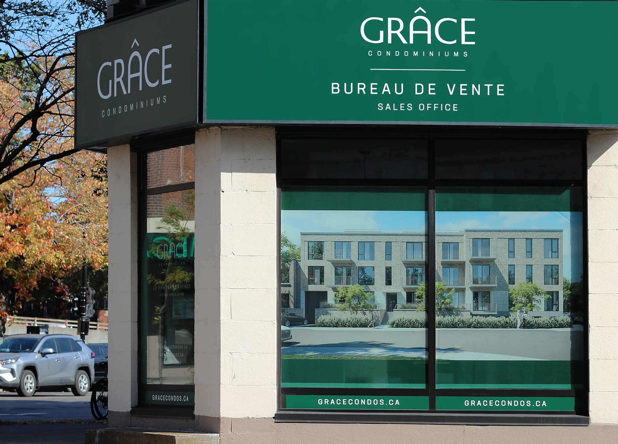 Grace_3 A sales office for Grace Condominiums with green signs in English and French, and a large window display showing a rendering of modern townhomes. Trees and parked cars are visible outside.