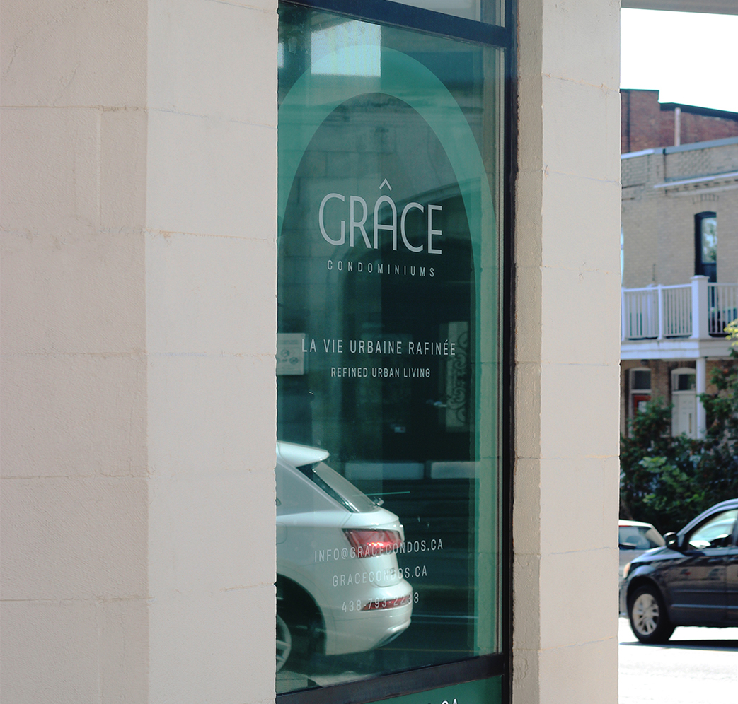 Grace_7 A window with a teal-green sign for Grace Condominiums, displaying the slogan “LA VIE URBAINE RAFFINÉE—Refined Urban Living,” along with contact information. A parked car and residential buildings are visible outside.