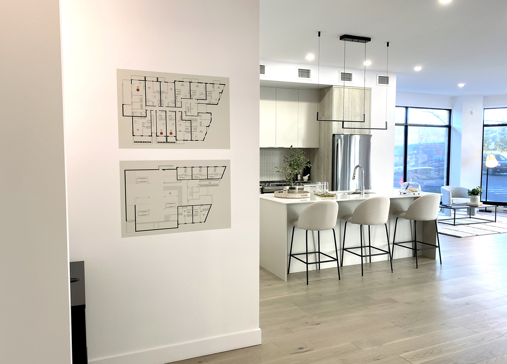 Grace_8 A modern, open-concept kitchen and living area with light wood floors, a central island with four stools, and large windows. Two floor plans are mounted on a white wall near the kitchen entrance.