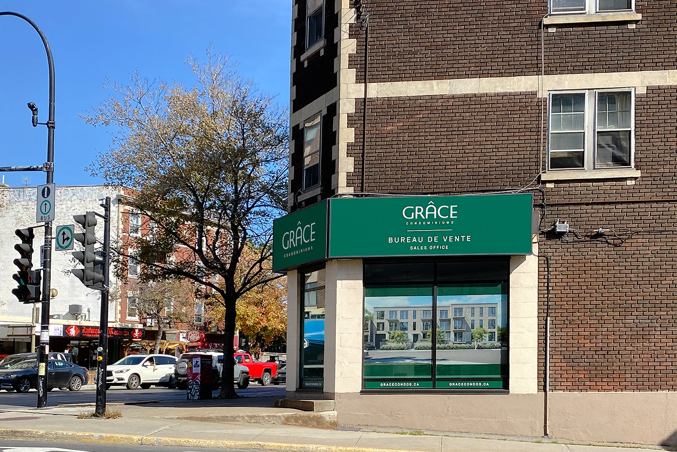 A street corner with a brick building featuring a green sign for "GRACE Condominiums" and "Bureau de Vente Sales Office." Large window displays condo images. Cars and pedestrians are visible; a tree stands nearby.