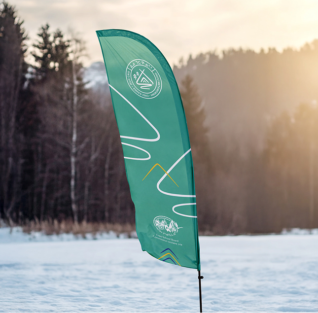 A green feather flag with white and yellow designs stands in snowy terrain, with trees and mountains in the background under a cloudy sky at sunset. The flag features circular logos and abstract line patterns.