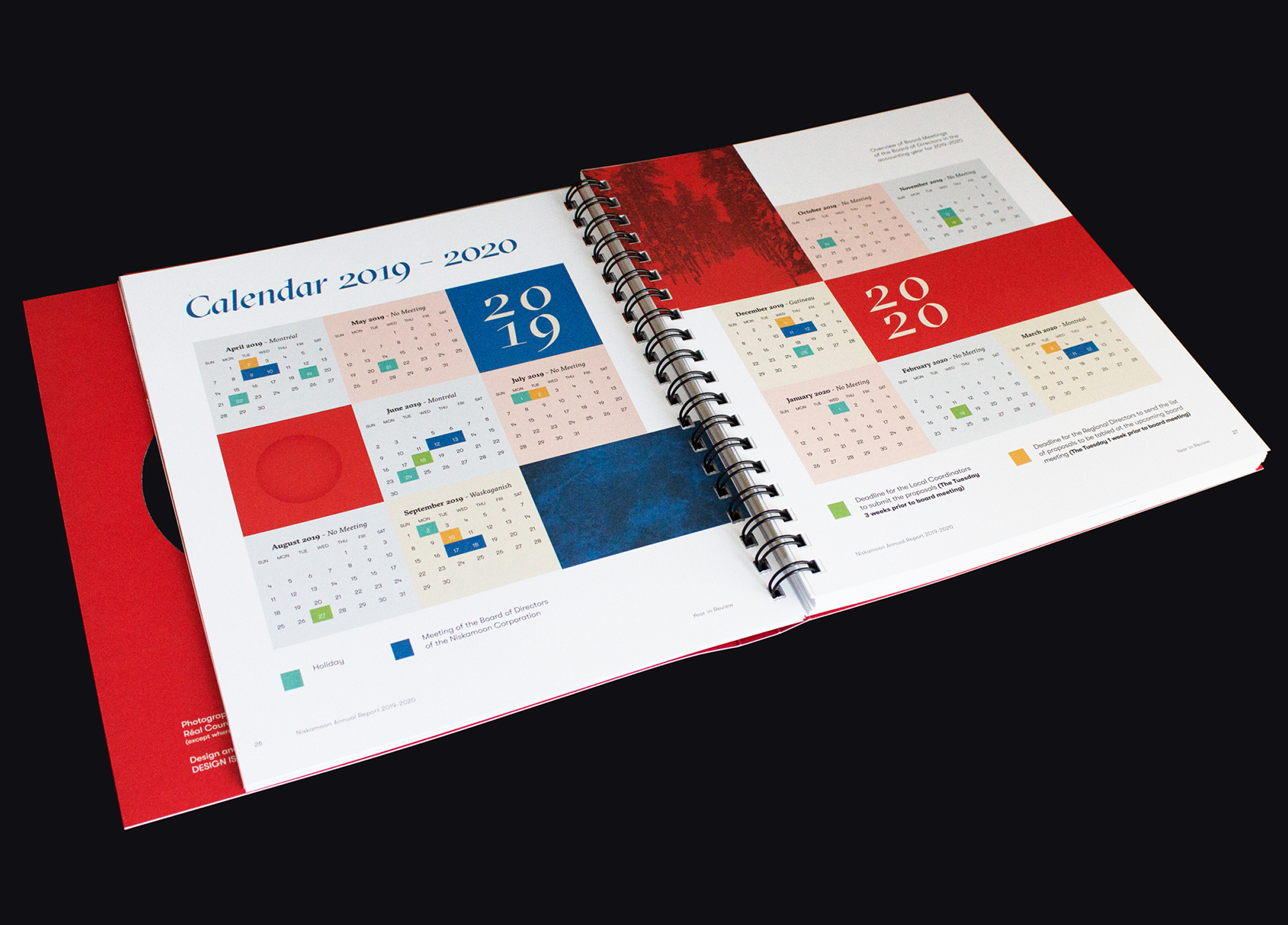Niskamoon_201920-4 An open spiral-bound planner displays a colorful calendar layout for 2019–2020, with months arranged in grids on both pages and different sections highlighted in various colors. The background is black.