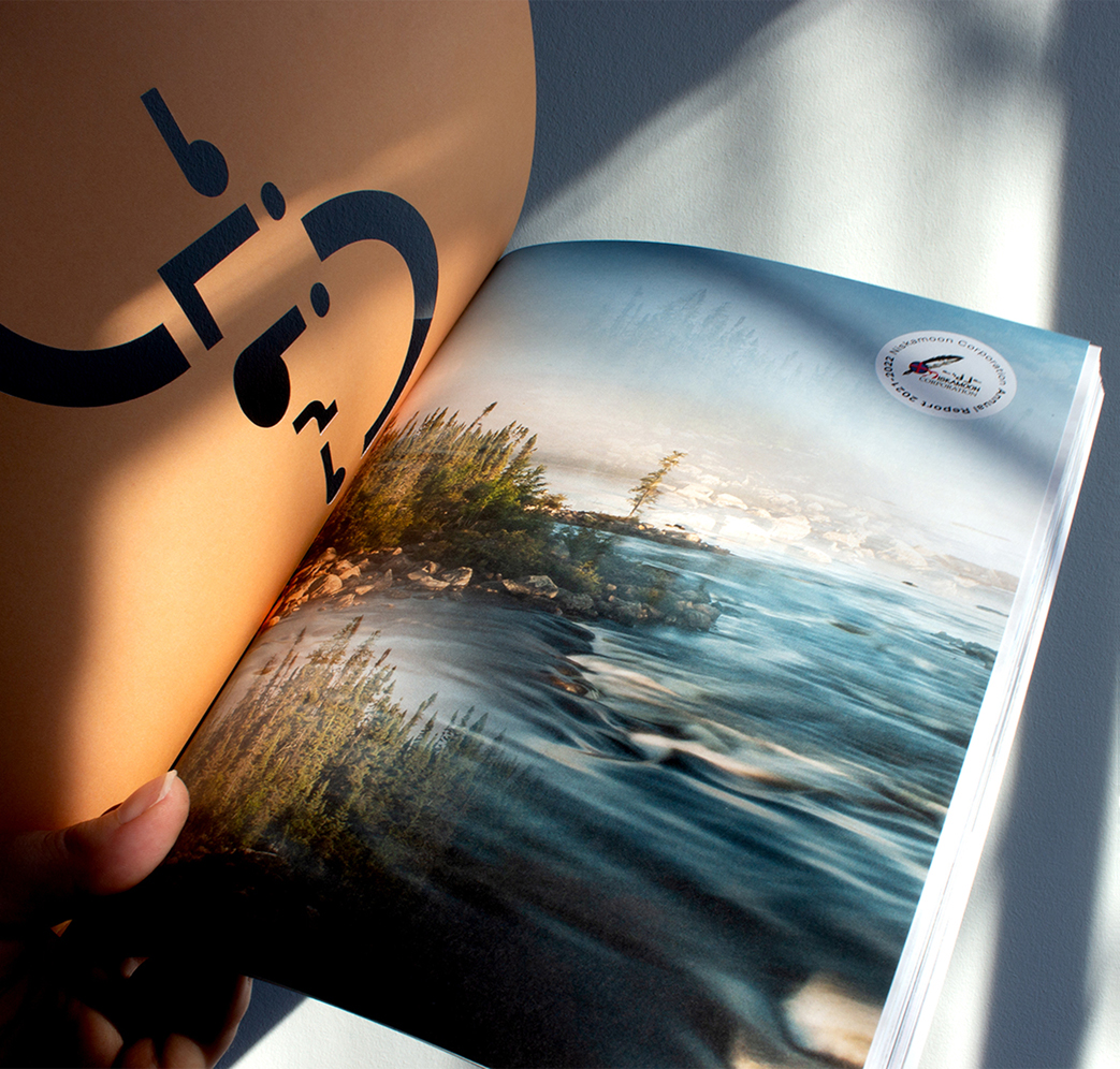 Niskamoon_202122-2 A hand holds open a magazine showing an artistic photo of a rushing river with trees alongside it, under sunlight. The left page shows abstract black shapes on a brown background.