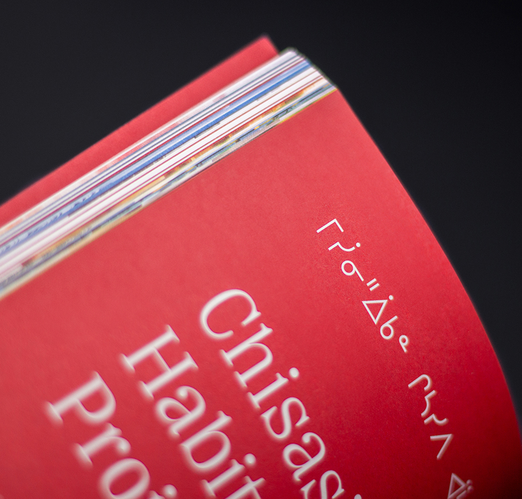 Niskamoon_202223-3 A close-up of a red book cover with white text in English and syllabic script, possibly Cree, against a black background. The visible English words include