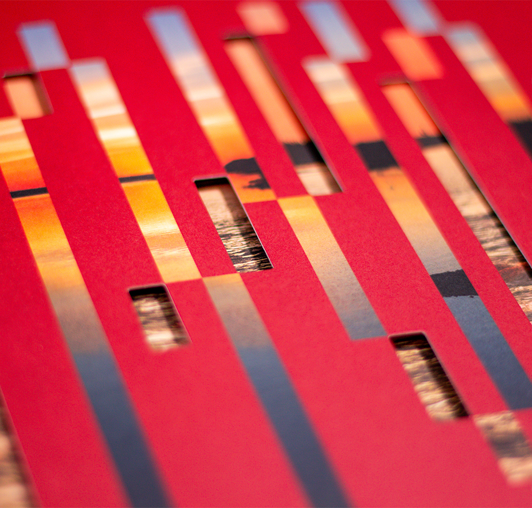 Niskamoon_202223-5 A close-up of a red surface with vertical rectangular cutouts, revealing parts of a sunset photograph with warm tones of orange and yellow over water beneath.