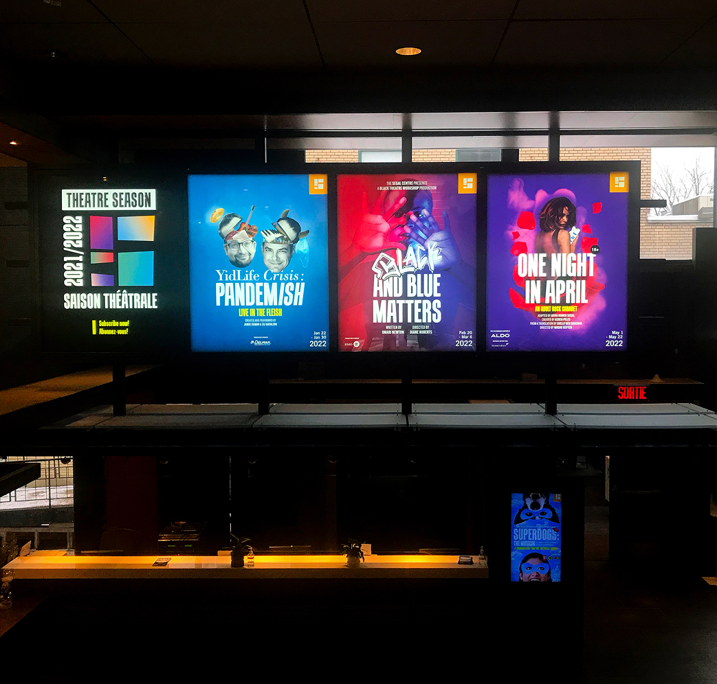 Segal_202122-2 Four colorful theater posters are displayed above a dark ticket counter. The posters promote different shows and the theater season, with bold titles and graphics, inside a modern building with large windows in the background.