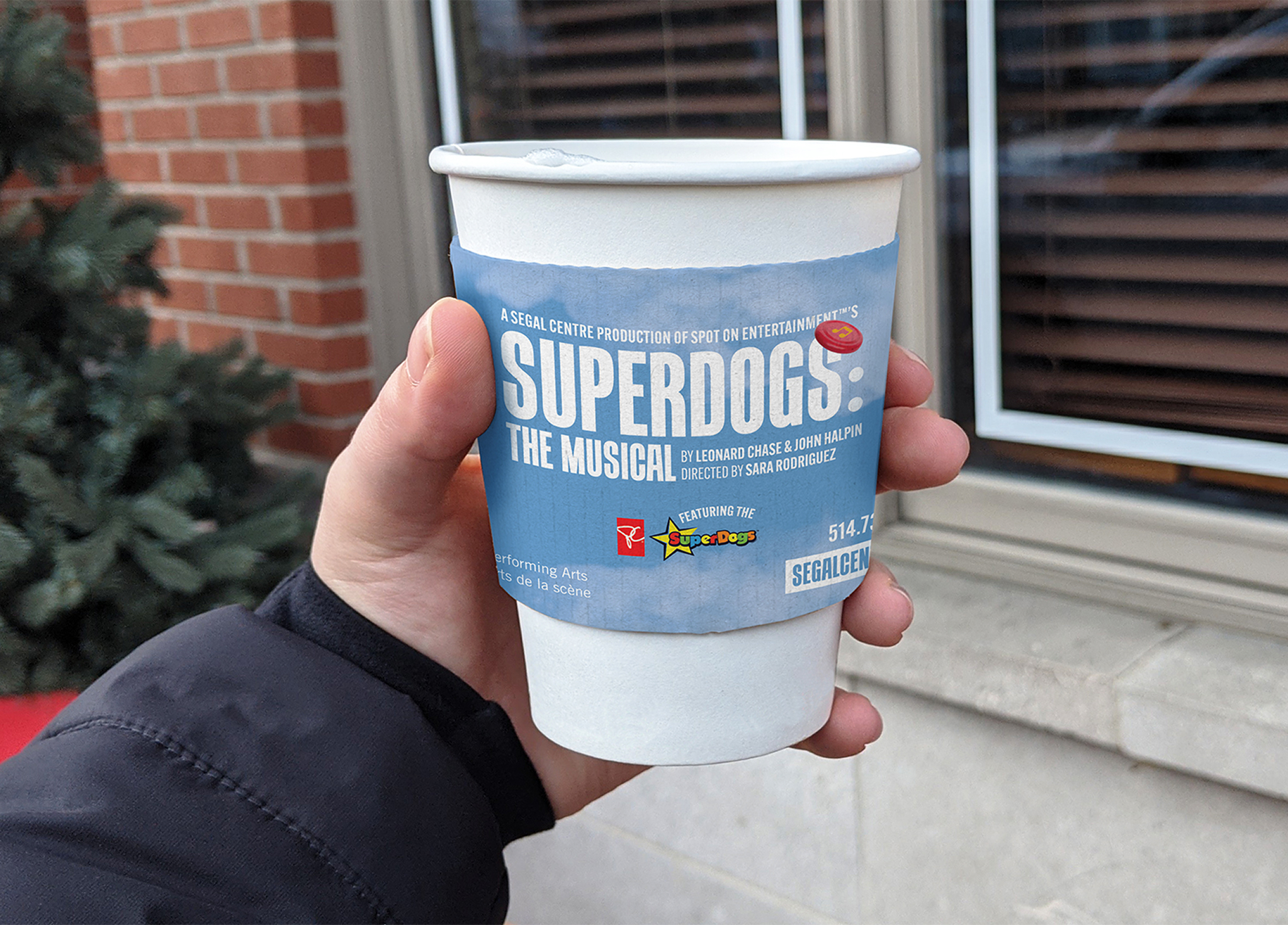 Segal_202122-7 A hand holds a white paper coffee cup wrapped in a blue sleeve that advertises