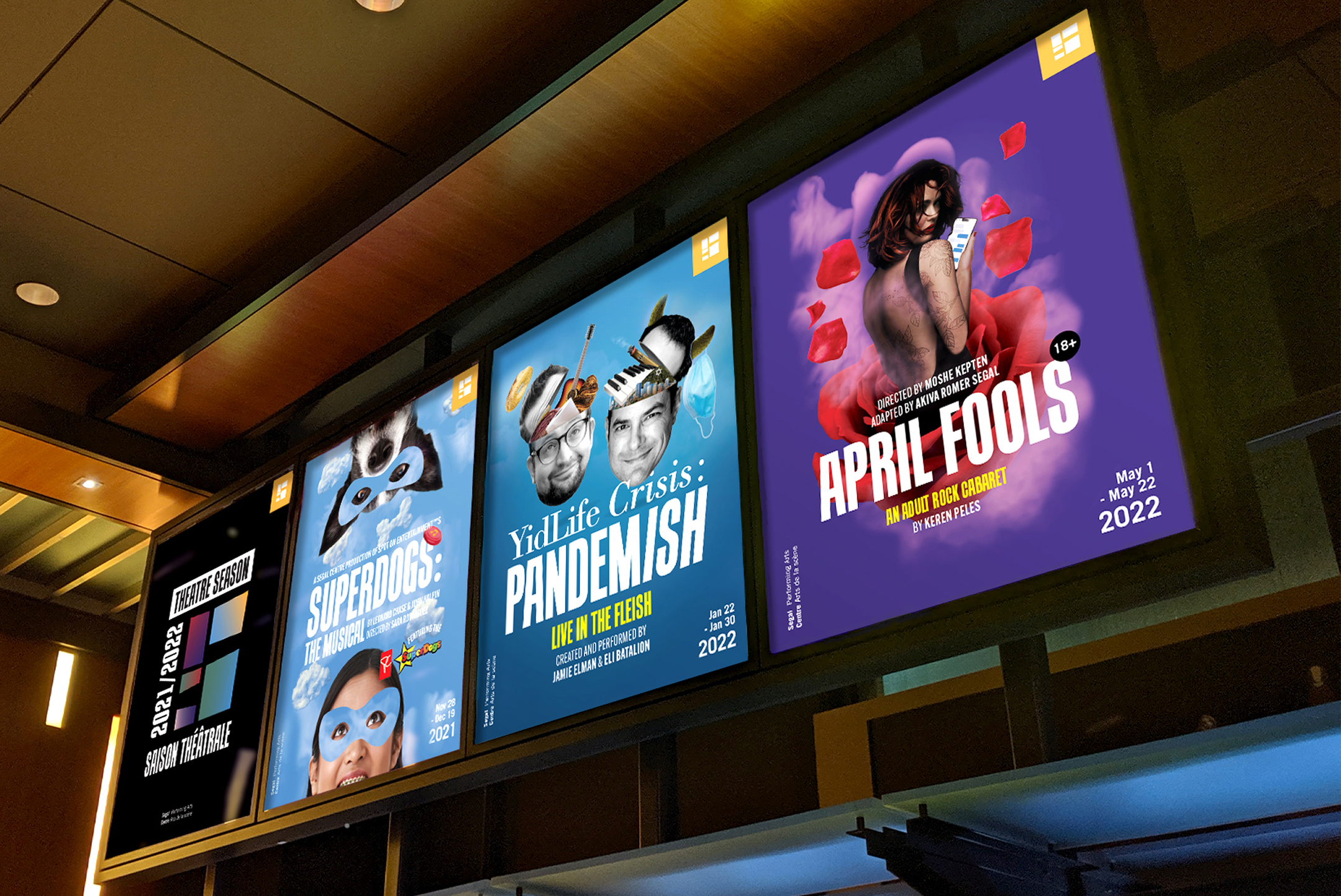 Three colorful theater posters on a wall display: “Superdog” with a superhero dog, “YidLife Crisis: Pandemish” featuring two men, and “April Fools” showing a surprised woman surrounded by red rose petals.