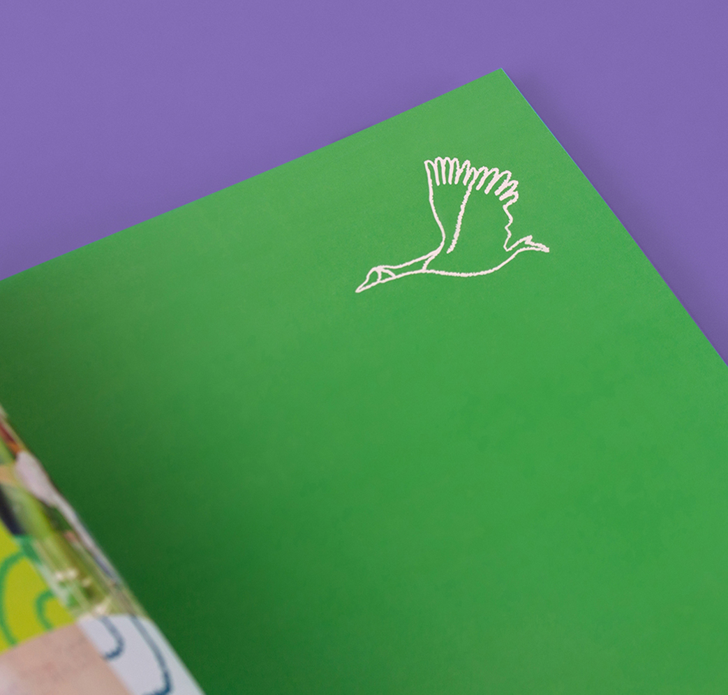 CSB_202324-2 An open book reveals a green page with a simple white outline drawing of a flying bird, likely a goose, in the upper right corner. The background is purple.