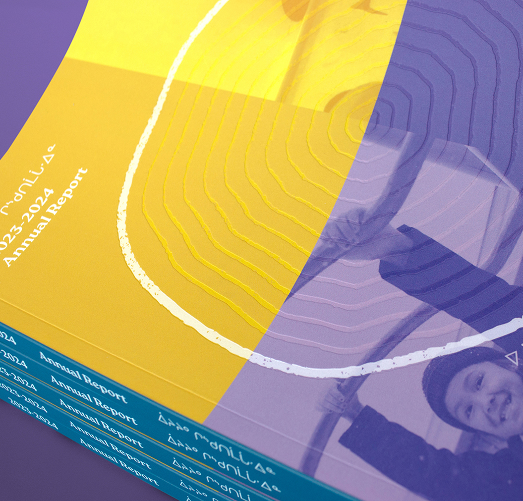 CSB_202324-5 Close-up of stacked annual reports with a vibrant cover design. The cover features abstract yellow and purple shapes, white contour lines, and a partial black-and-white photo of a smiling person holding a hoop.