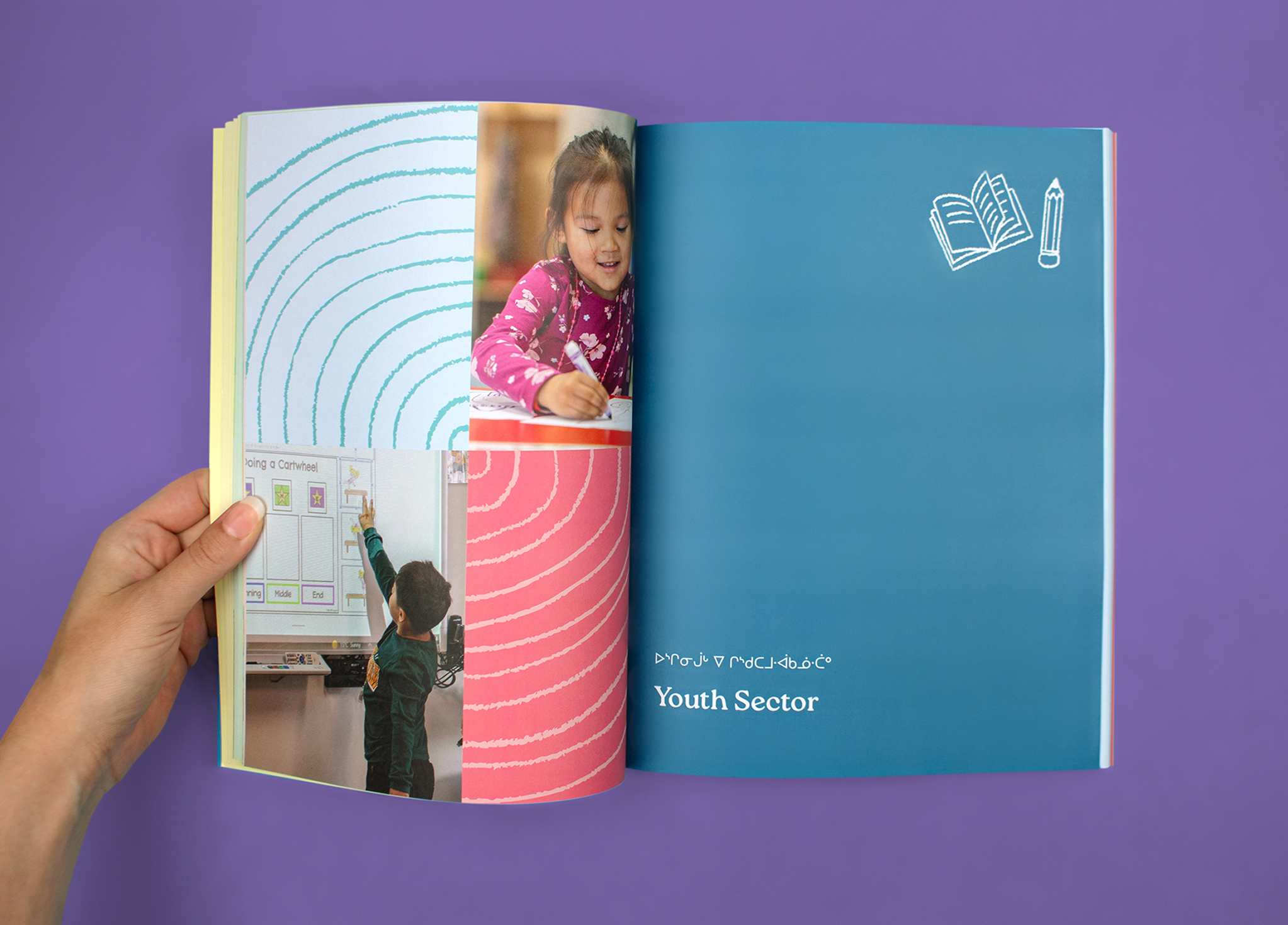 CSB_202324-6 A hand holds open a colorful book. On the left page, there are photos of a child writing and another pointing at a board, with wavy lines. The right page is blue with white drawings of a book, pencil, and text