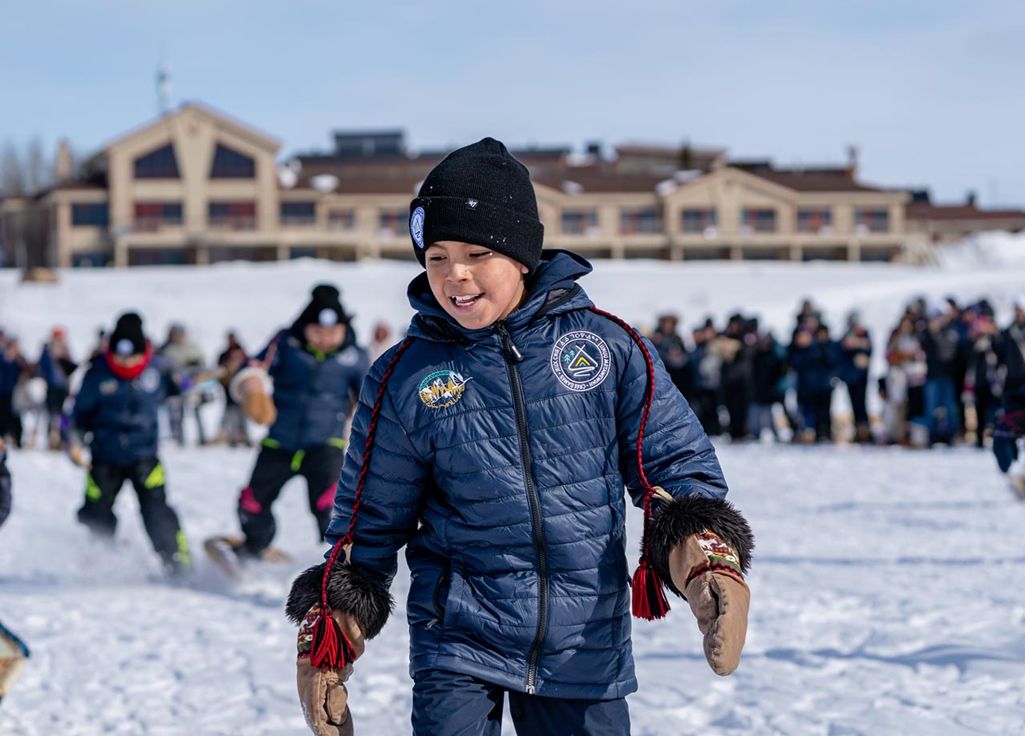 A smiling child in a blue winter coat, hat, and mittens runs on snow, with other children and a crowd in the background near a large building on a sunny winter day.