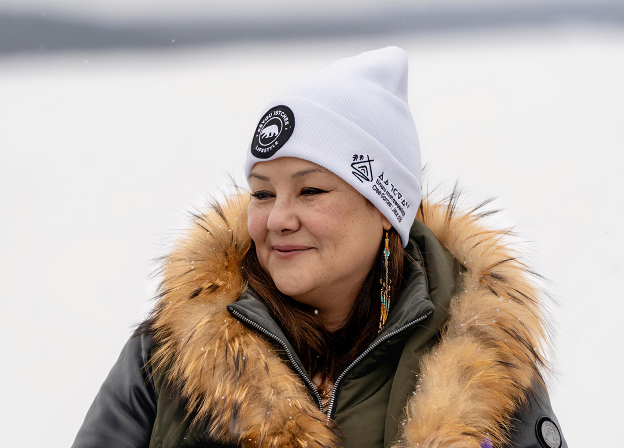 A woman wearing a white beanie with logos, a fur-lined hooded jacket, and beaded earrings stands outside in a snowy landscape, smiling gently.