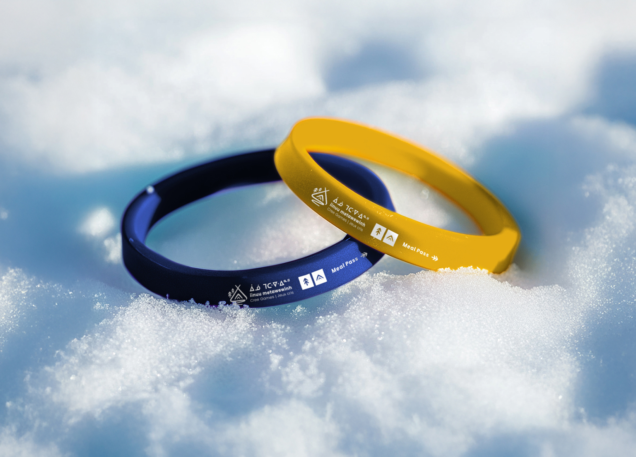 Two silicone wristbands, one blue and one yellow, rest on a bed of snow. Both bands have white printed mountain graphics, symbols, and some text on them. The scene suggests a cool, outdoor environment.