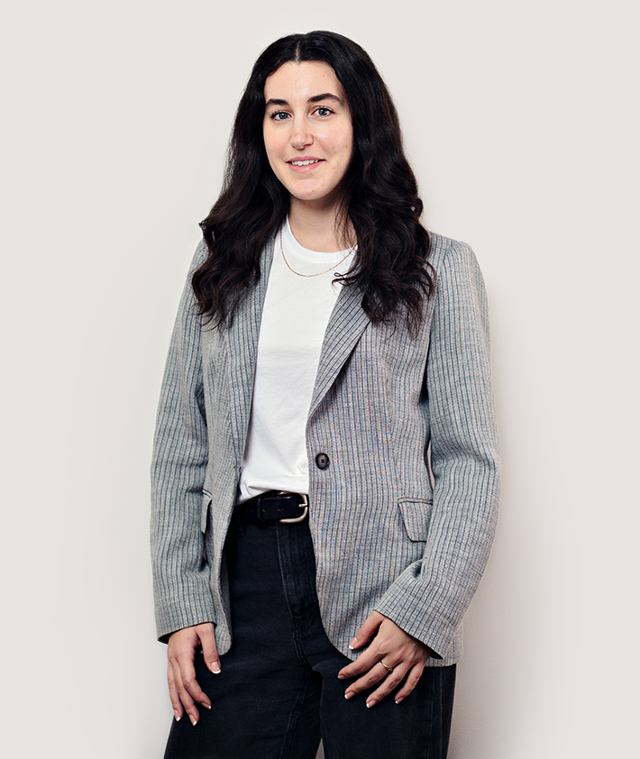 A woman with long dark hair wearing a gray striped blazer over a white shirt and black pants stands smiling against a plain light background.